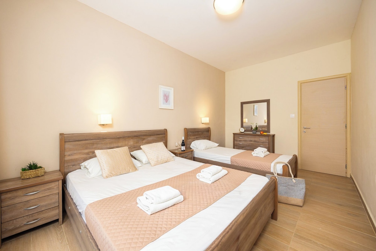 A hotel room with two beds, wooden headboards, bedside tables with lamps, a mirror, and a door.