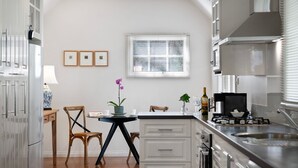 A modern kitchen with white cabinetry, a dining area with a table and chairs, and a window with blinds. - Haiku- charming oasis in Daylesford (Daylesford)