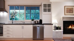 A modern kitchen with white cabinetry, a dishwasher, a window with a view of greenery, a fireplace, and a dining area with a table and chairs. - Haiku- charming oasis in Daylesford (Daylesford)