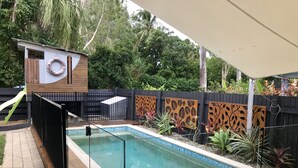 Piscina retangular com borda revestida em azulejo, rodeada por um deck de madeira. Há uma cerca preta com painéis decorativos e uma pequena construção de madeira com uma placa circular.
