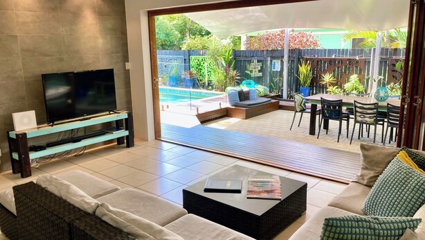 Uma sala de estar moderna com televisão de tela plana, sofá, mesa de centro e porta de vidro deslizante que dá para um pátio externo com piscina e área de estar.