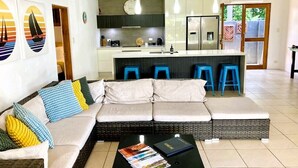 Uma sala de estar com sofá, mesa de centro e área de cozinha com geladeira e banquetas.