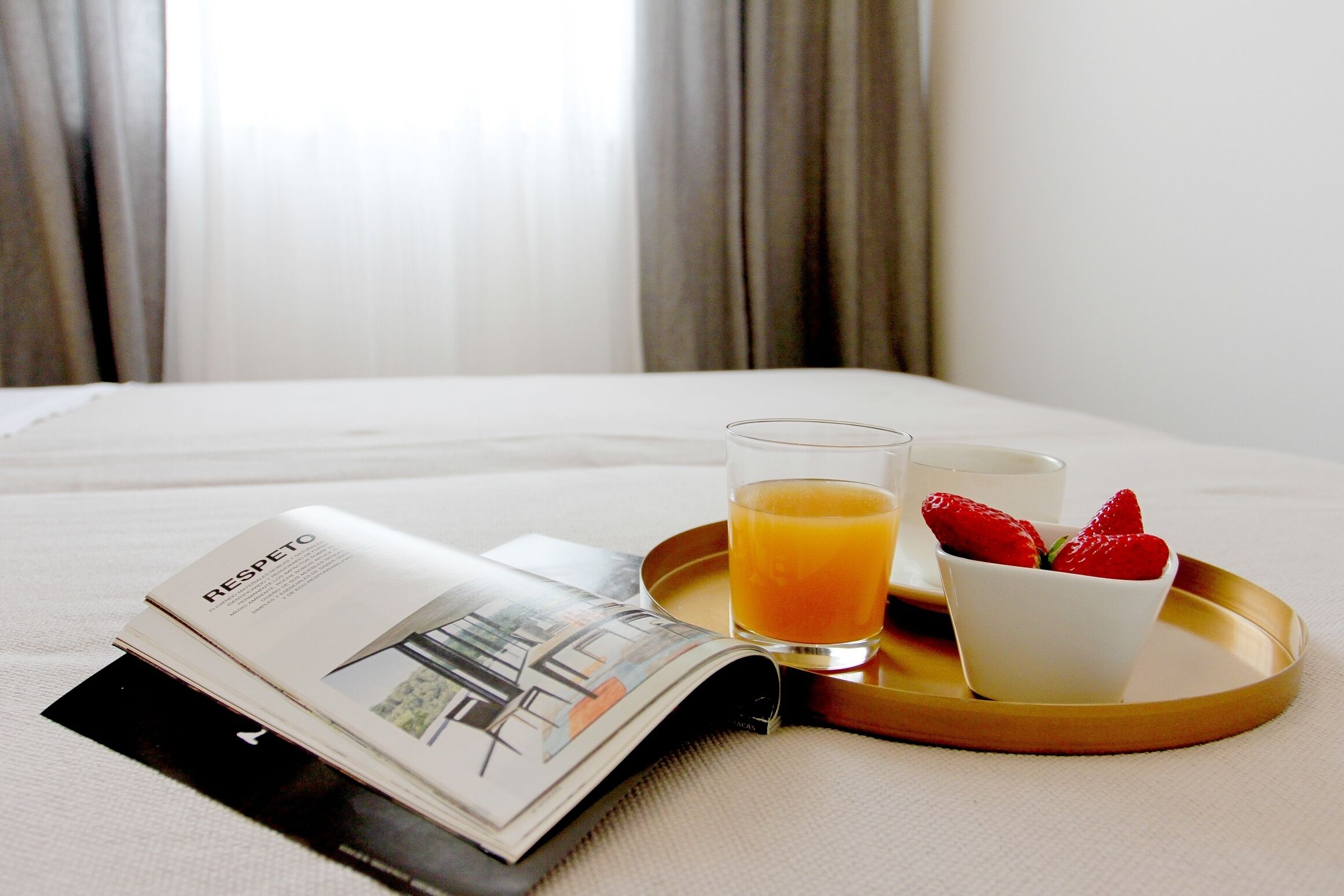 A neatly made bed with a tray containing a glass of orange juice, a cup, and a bowl of strawberries.