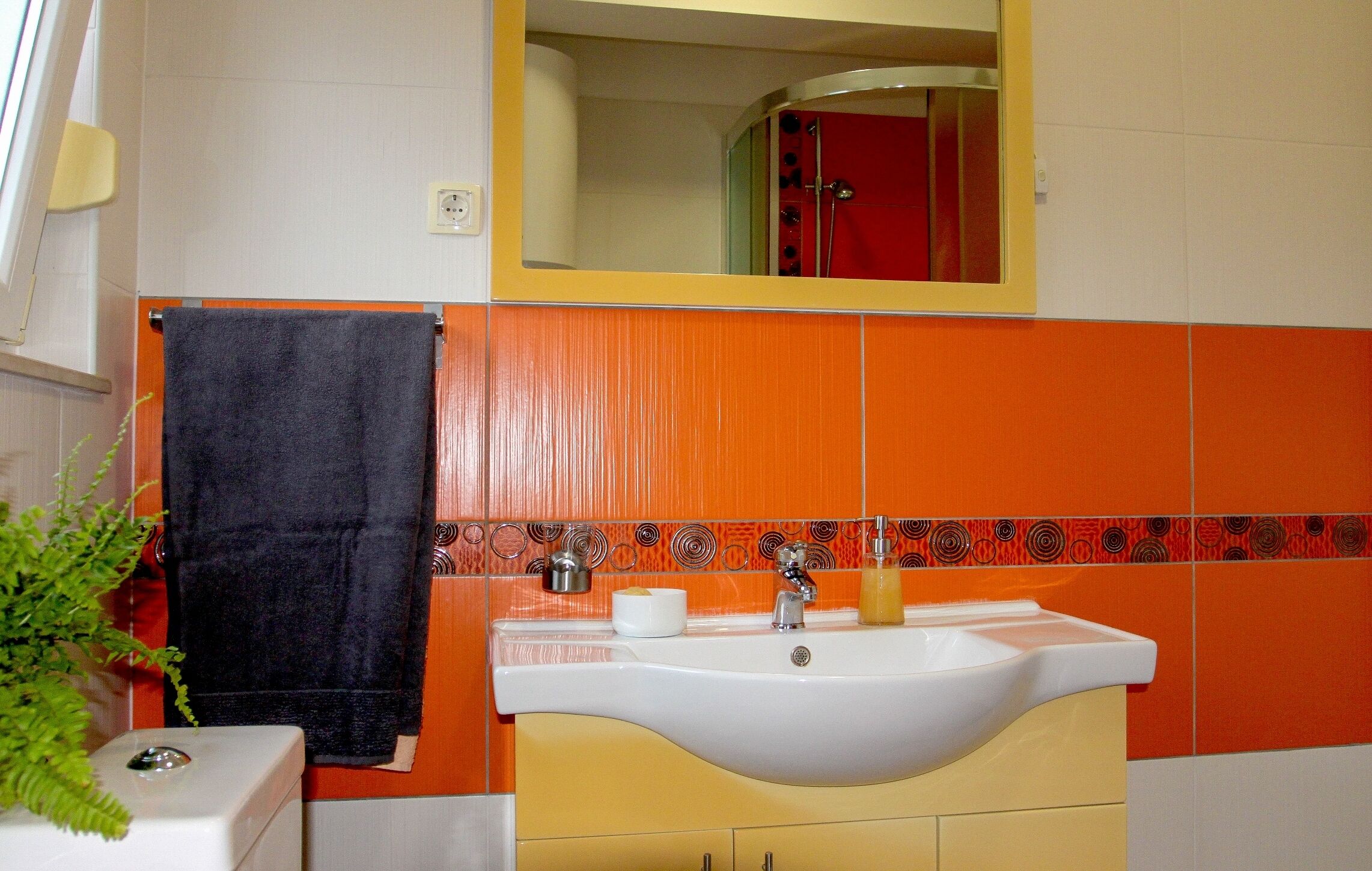 A bathroom with a white sink, orange and white tiles, a black towel, and a mirror above the sink.