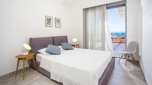 Un dormitorio con cama, dos mesitas de noche, una silla, un balcón con vistas y dos cuadros en la pared.