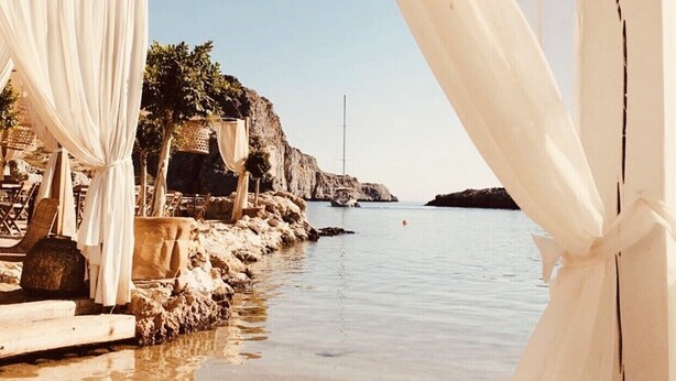 Una playa con cortinas blancas, un litoral rocoso y un velero a lo lejos.