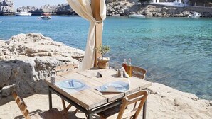 Una mesa de madera con un mantel blanco, una lámpara de mimbre y una pequeña planta en maceta sobre un paisaje rocoso junto al mar.