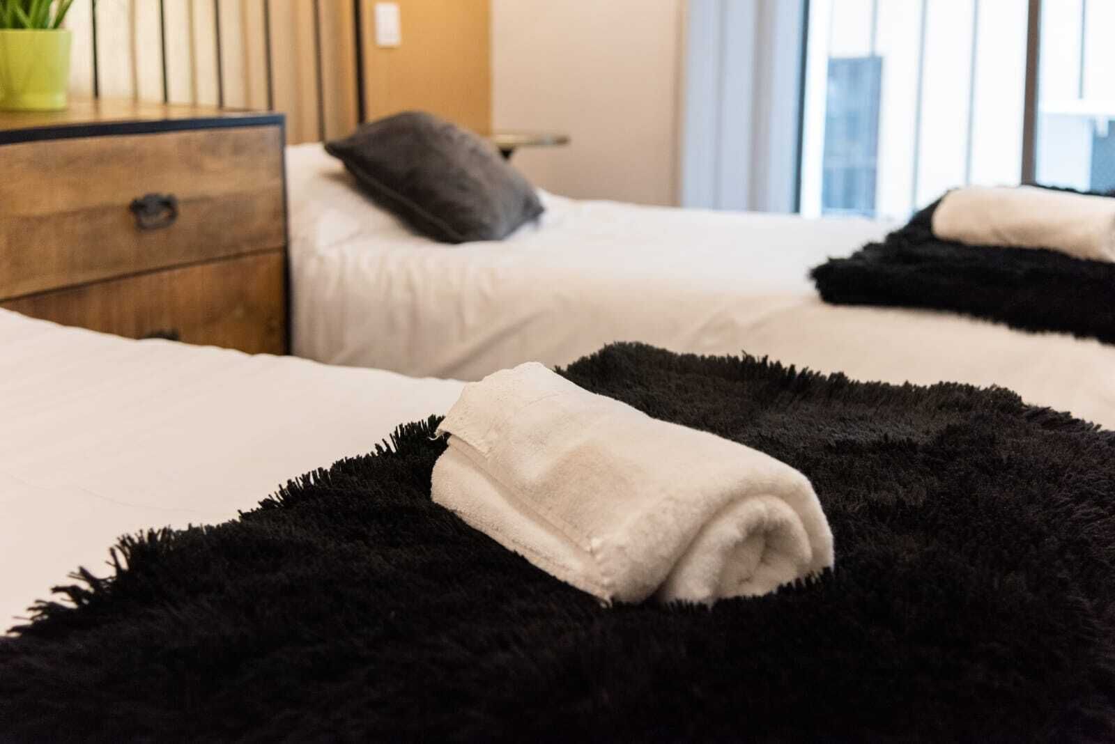 A hotel room with two beds, a wooden nightstand, a green plant, and a window with blinds.