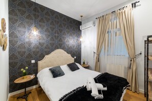 A bedroom with a patterned wall, a bed with a white and black bedspread, two pillows, a bedside table with a plant, and a hanging towel.
