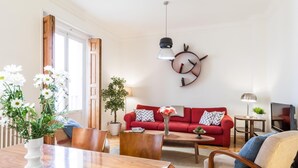 A living room with a red sofa, wooden coffee table, and a dining table with chairs. There is a potted plant and a wall clock.