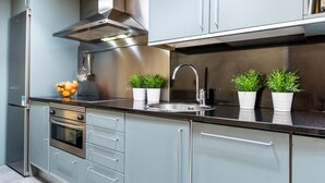 A modern kitchen with stainless steel appliances, a built-in oven, and a sink with a faucet. There are potted plants on the countertop and a bowl of fruit on the left.