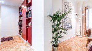 A spacious room with a herringbone wooden floor, a red bookshelf filled with books, a potted plant, and a framed abstract artwork on the wall.