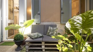 Uma área externa com assento acolchoado, uma planta em vaso e uma porta que dá para um quarto.