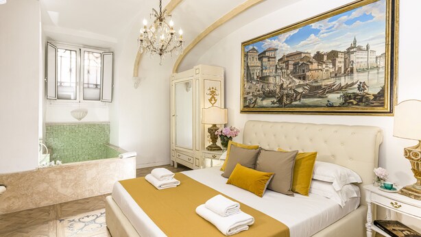 Quarto de hotel com uma cama grande, banheira, candelabro, pintura e espelho.