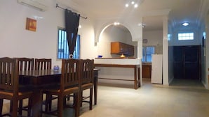Een ruime eetruimte met een houten tafel en stoelen, een keuken op de achtergrond, en uitzicht buiten door een raam.