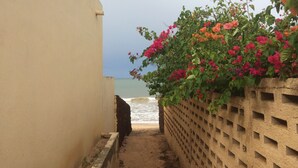 Een smal pad dat naar een strand leidt, met aan één kant een betonnen muur en aan de andere kant een gebouw, overwoekerd met bloeiende bougainvillea.