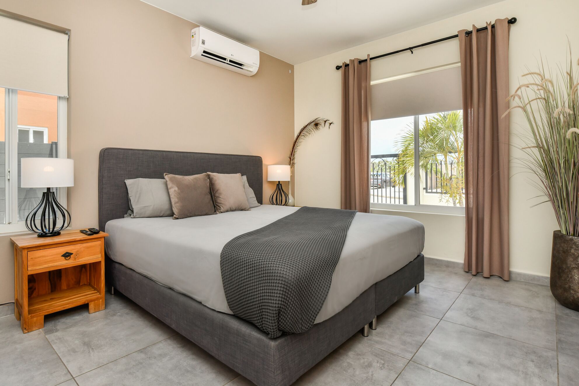 Una habitación de hotel moderna con cama, mesita de noche, lámpara, unidad de aire acondicionado, ventana con cortinas y vistas a palmeras.