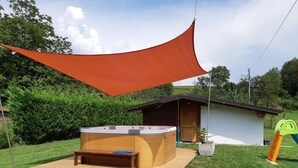 Un bain à remous installé sur une plateforme en bois, avec un banc en bois, un petit abri et une structure de jeux dans une zone herbeuse, avec des arbres en arrière-plan.