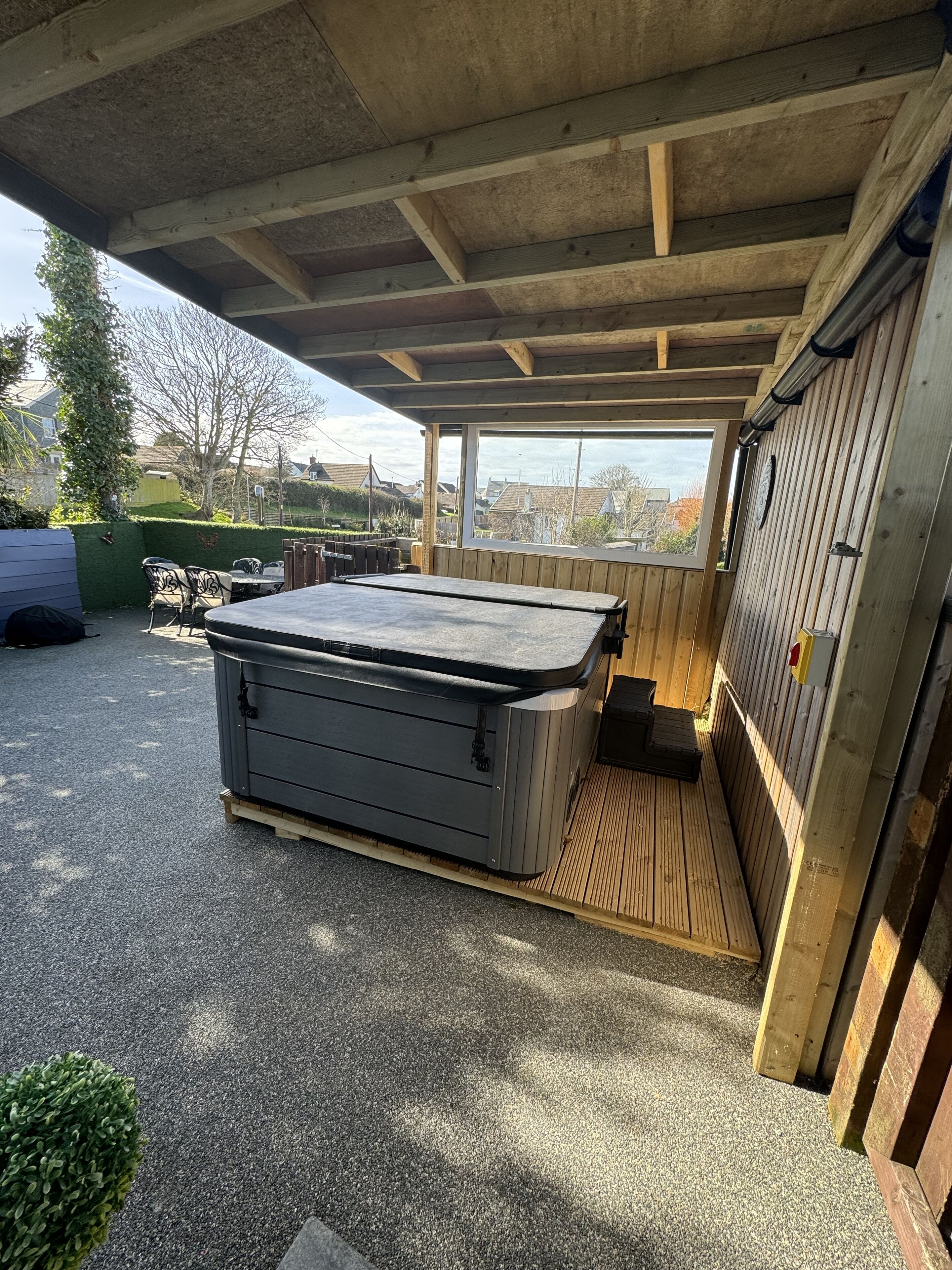 Een overdekt terras buiten met een hottub, een houten terras en uitzicht op de omgeving.