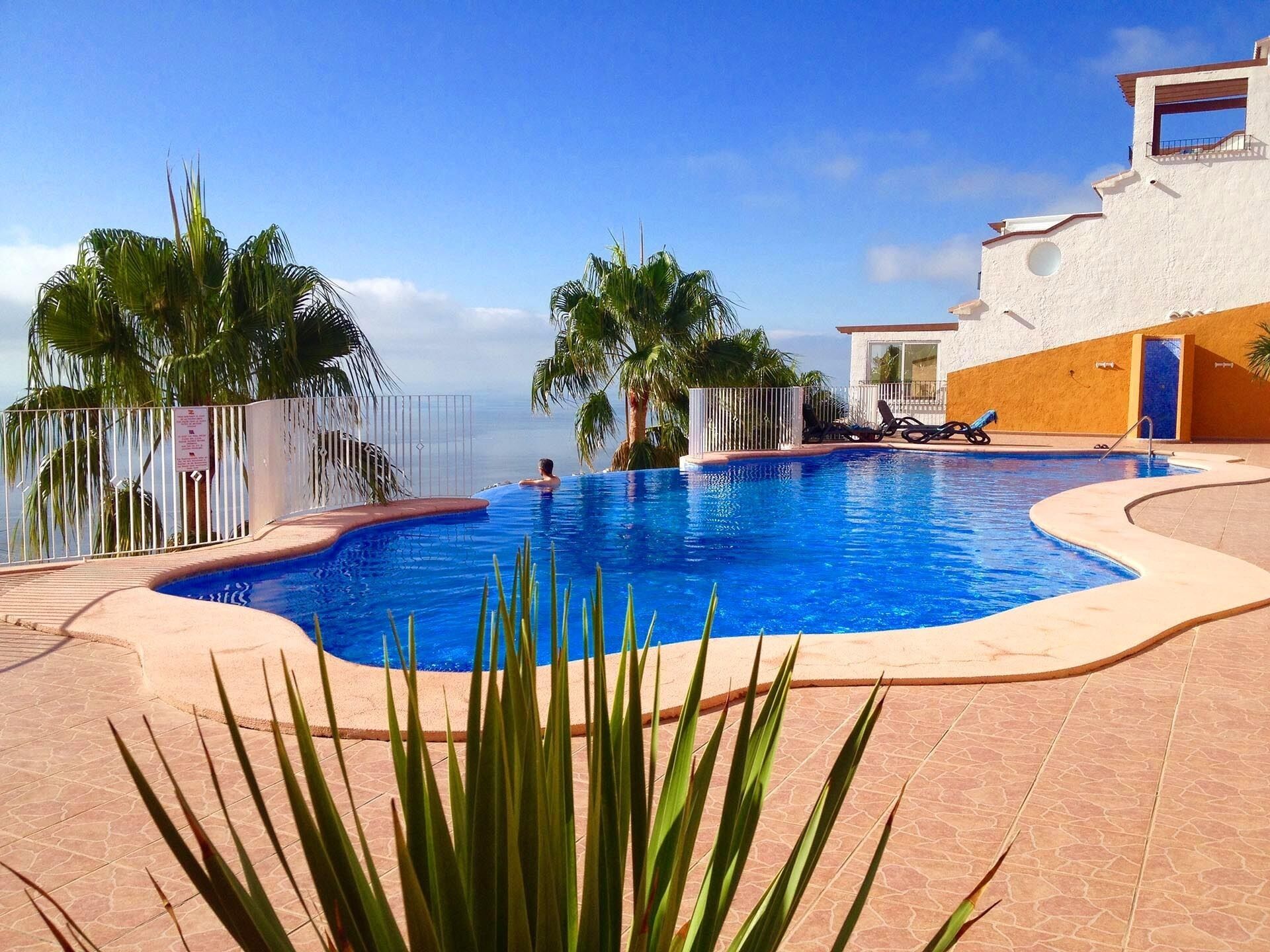 A swimming pool with a view of the ocean, surrounded by palm trees and a white building with a balcony.