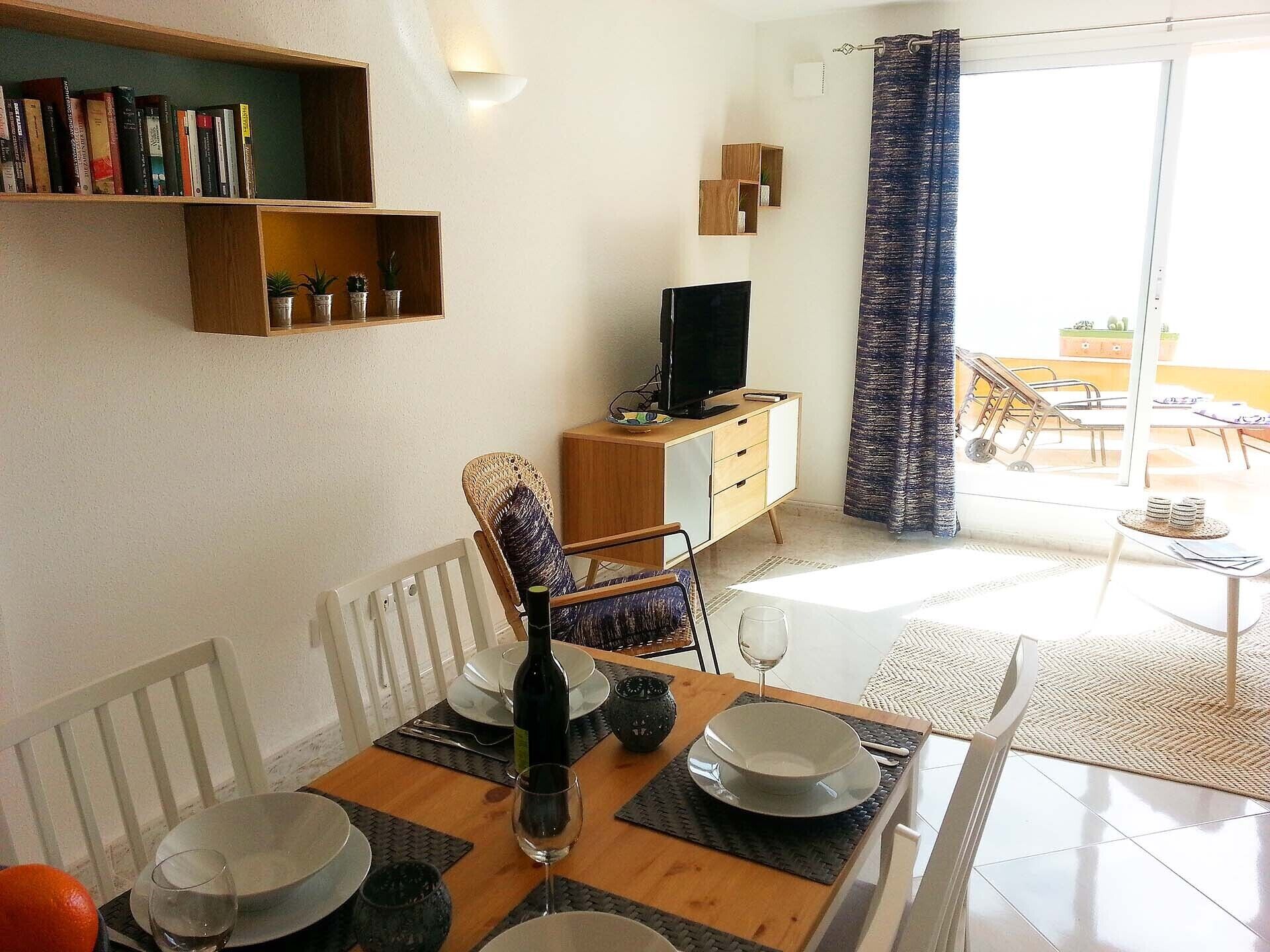 A dining area with a wooden table set for two, a television on a wooden cabinet, and a balcony with a view.
