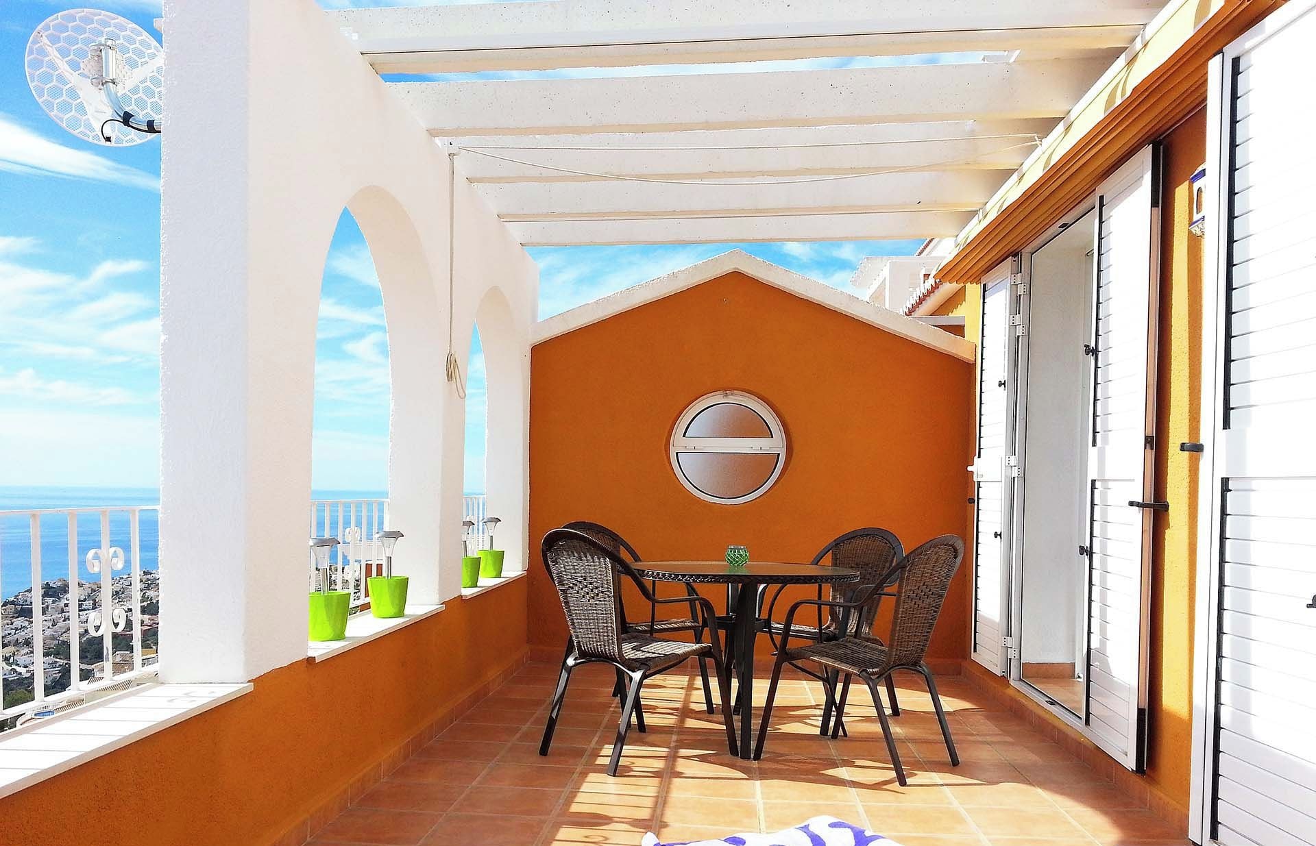 A balcony with a table and chairs, a view of the sea, and a white wall with a window.