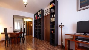 A living room with a wooden floor, a dining table with chairs, a bookshelf, a television, and a lamp.