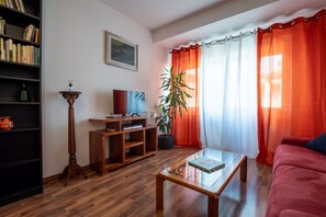 A living room with a wooden floor, a red sofa, a coffee table, a TV on a wooden stand, a bookshelf, a potted plant, and orange curtains. - Rialto Apartment Vatican Museums (Rome)