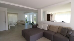 A modern living room with a large sectional sofa, a white armchair, and a glass coffee table. There is a large window with curtains, a cabinet, and a mirror on the wall. - Deluxe Villa with private pool and jacuzzi 2km from Ortigia, Sicily (Siracusa)