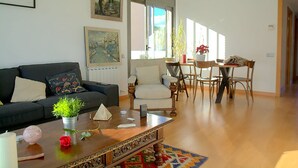 Sala de estar com sofá, mesa de centro e cadeiras. Há quadros nas paredes e uma janela com cortinas.