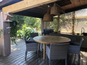 Een terras buiten met een ronde houten tafel en rieten stoelen, een hanglamp en uitzicht op groen door een raam.