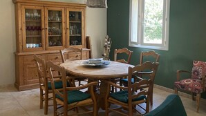 A dining area with a round wooden table, six chairs, a wooden cabinet with glass doors, and a window with a view of greenery. - 5 bedroom VILLA with pool on a hillside near AIX  (Mimet)