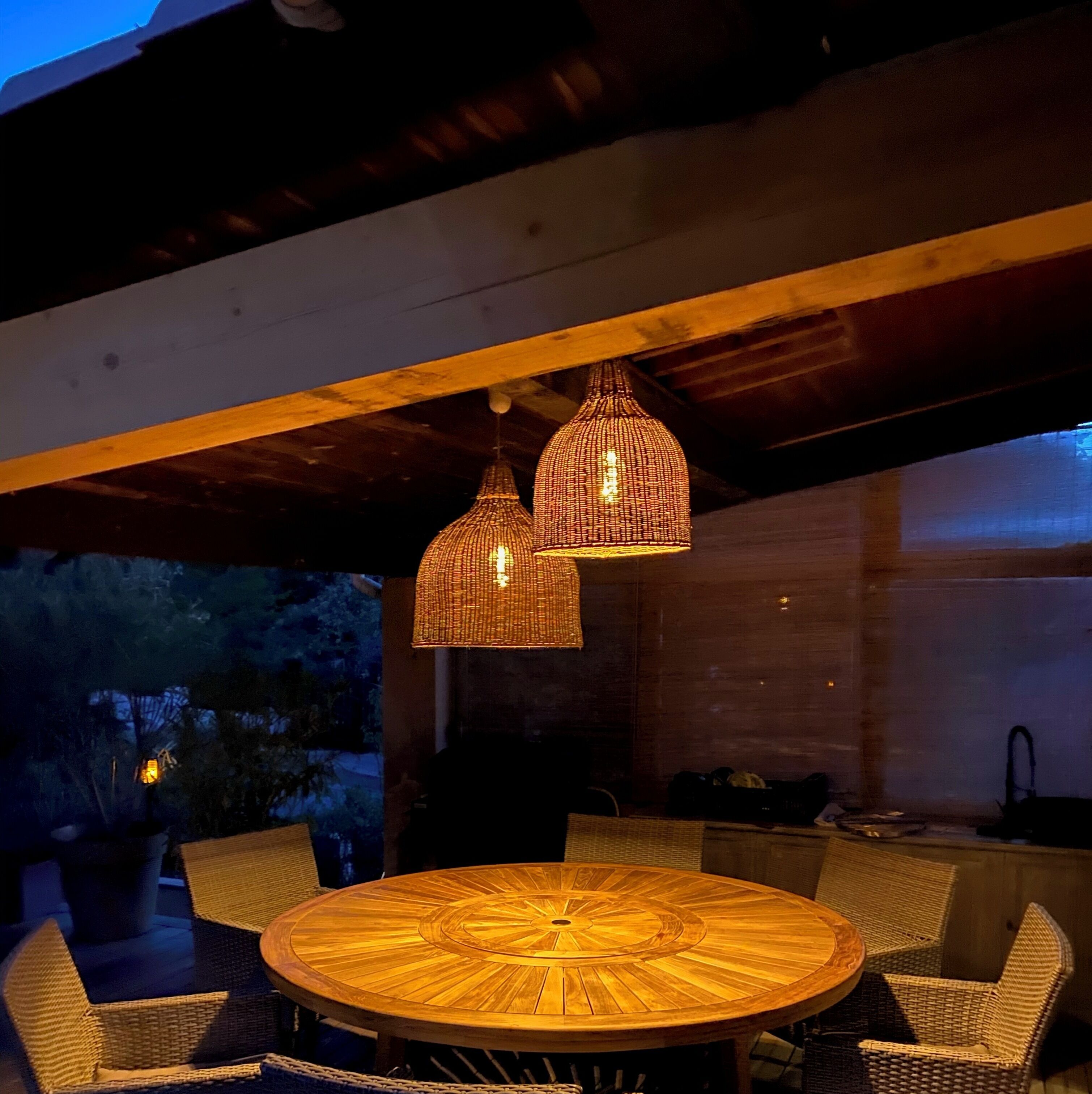 Une table ronde en bois, entourée de chaises en osier, deux lampes suspendues et une vue sur l’extérieur.