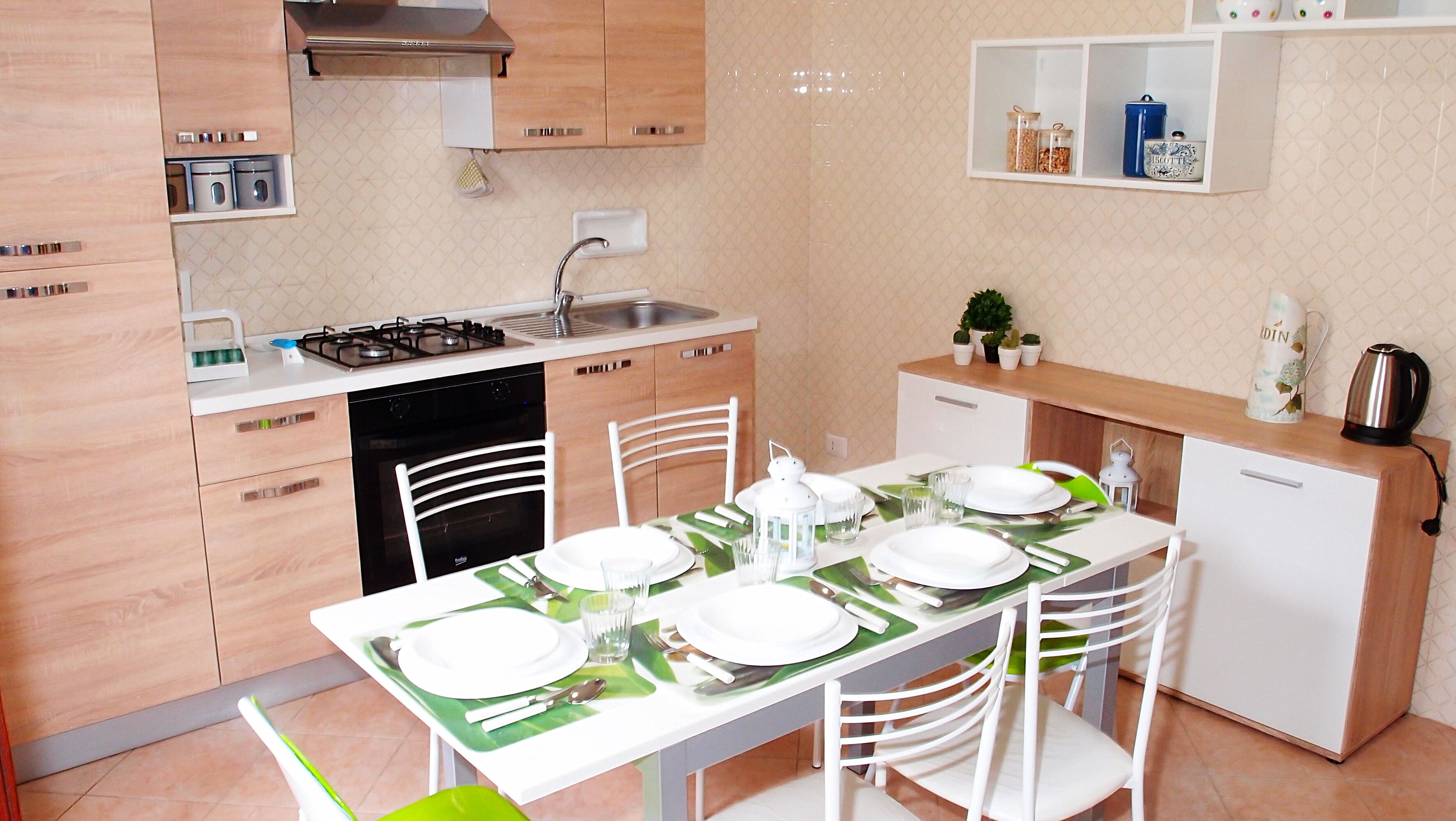 Une cuisine moderne avec des meubles blancs, une cuisinière à gaz, un lave-vaisselle et une table à manger pour quatre personnes, avec de l’assiette blanche et des sets de table verts.
