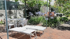 A patio with white lounge chairs and a table, surrounded by greenery and a brick wall.