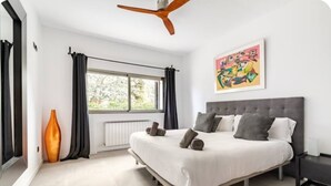 A modern bedroom with a large bed, a ceiling fan, a window with curtains, and a painting on the wall.