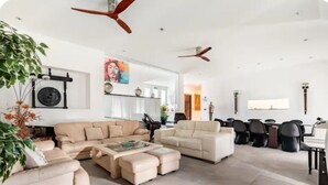 A spacious living room with a large L-shaped sofa, a smaller sofa, and a coffee table. There are ceiling fans, a dining area with chairs, and a large window providing natural light.