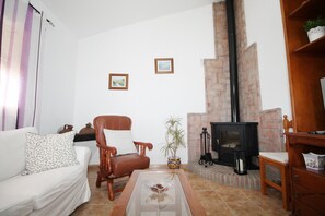 Un salon avec un canapé blanc, un fauteuil en bois, une table basse en verre, une cheminée et deux tableaux encadrés accrochés au mur.