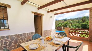 A dining area with a table set for four, stone walls, a wooden door, and a view of a hilly landscape. - 1076 Cortijo de la Musica (Frigiliana)