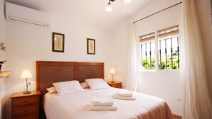 A bedroom with a wooden headboard, two bedside tables with lamps, a bed with a patterned bedspread, and a window with blinds. - 1076 Cortijo de la Musica (Frigiliana)