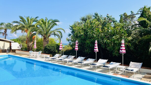 Una piscina con sillas de descanso y sombrillas a rayas, rodeada de palmeras y vegetación.