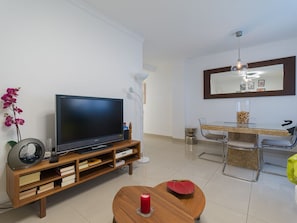 Uma sala de estar moderna com uma TV de tela plana sobre um suporte de madeira, uma mesa de jantar com cadeiras e um espelho grande acima da mesa.
