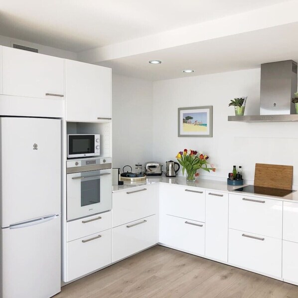 A modern kitchen with white cabinetry, a built-in oven, microwave, and a toaster. There is a wooden cutting board on the countertop and a vase with flowers. - Modern House With Panoramic Sea Views in Mallorca (Son Serra de Marina)