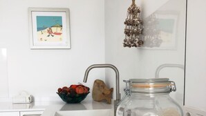 A kitchen with a white countertop, a sink, a bowl of fruit, and a decorative jar with seashells and blue beads.
