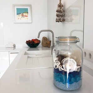 A kitchen with a white countertop, a sink, a bowl of fruit, and a decorative jar with seashells and blue beads. - Modern House With Panoramic Sea Views in Mallorca (Son Serra de Marina)
