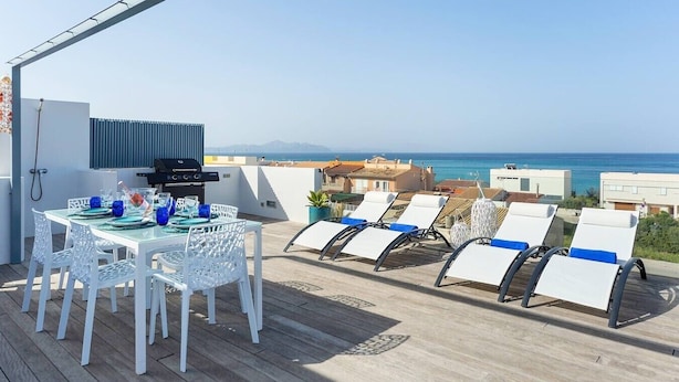 Une terrasse sur le toit avec une table à manger, des chaises, un barbecue et des fauteuils, le tout offrant une vue sur la mer.