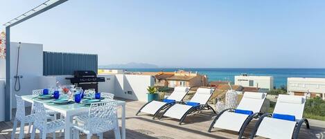A rooftop terrace with a dining table, chairs, a grill, and lounge chairs overlooking the sea.