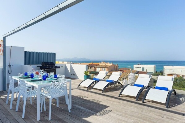 A rooftop terrace with a dining table, chairs, a grill, and lounge chairs overlooking the sea.