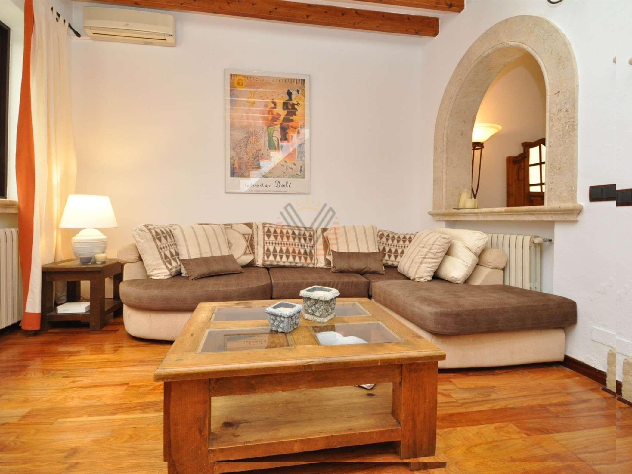 A living room with a wooden coffee table, a brown sofa with patterned cushions, a framed picture on the wall, and a stone archway.