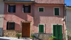 Ein zweistöckiges Gebäude mit steinernem Sockel und rosafarbenen oberen Wänden, das eine mittige Holztür und grüne Fensterläden aufweist.